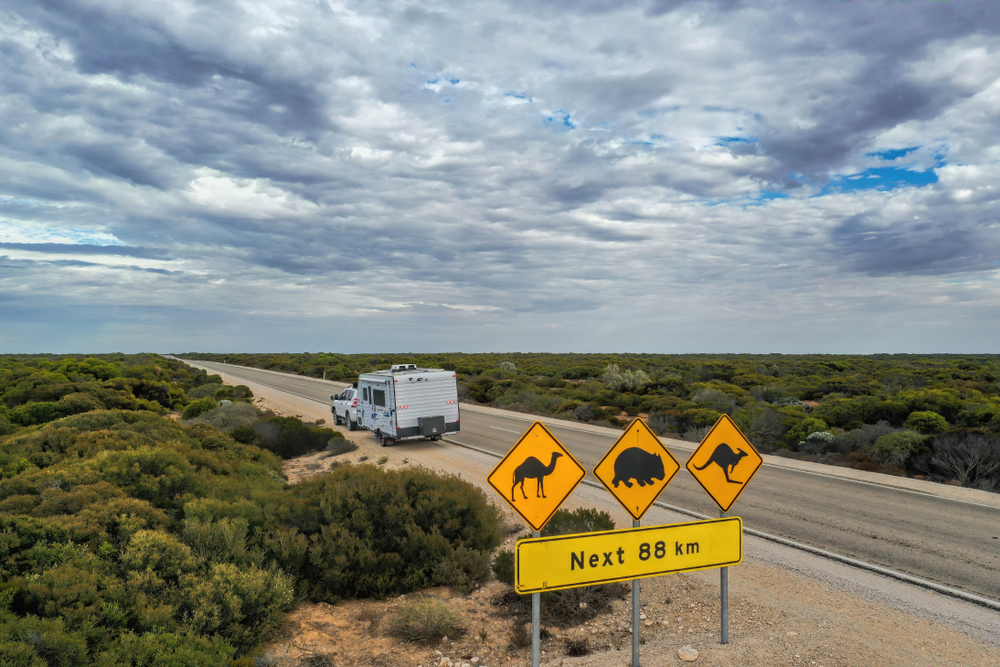A caravan being towed by a car travels on a road past some yellow warning signs