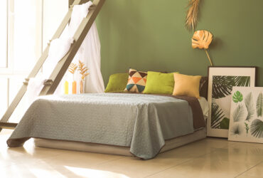 Large bed with against a green wall with colourful pillows and paintings on the ground